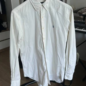 Vit skjorta från Ralph Lauren - Klassisk vit skjorta från Ralph Lauren med button down-krage och broderad blå logga på bröstet. Skjortan har lång ärm, knappar framtill och är tillverkad i bomull. Perfekt för en clean och stilren look.