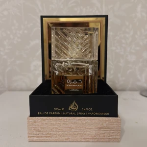 Khamrah parfym från Lattafa 70-75ml - Elegant parfymflaska i genomskinligt glas med dekorativa snitt och guldig etikett. Innehållet är bärnstensfärgat och flaskan har en fyrkantig form med matchande lock. Märkt med 'Khamrah' och 'Lattafa' på framsidan.