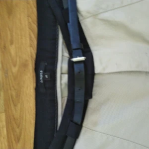 Beiga kostymbyxor från Lindex med bälte - Stilrena beiga kostymbyxor från Lindex i storlek 44. Byxorna har svarta detaljer i midjan och kommer med ett matchande svart bälte. Perfekta för en clean och klassisk look. Tillverkade i ett mjukt bomullsmaterial för skön känsla.