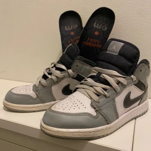 Nike Air Jordan 1 Mid Grå/Vit EUR 34 - Snygga Nike Air Jordan 1 Mid sneakers i grått och vitt med svarta detaljer och klassisk swoosh. Skorna har snörning, rund tå och platt sula. Tillverkade i skinn och syntetmaterial, perfekta för dig som gillar streetstyle och ikoniska sneakers. Hittade tyvärr inte den originala sulan med man får den Artapidrriska sulan med den för 50kr extra.