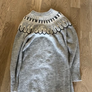 Grå stickad tröja från Vero Moda - Mysig grå stickad tröja från Vero Moda med snyggt mönster i vitt och svart upptill. Tröjan har lång ärm, ribbade muddar och en klassisk rund halsringning. Perfekt för kyliga dagar när du vill vara både varm och stilren.