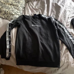 Svart Champion sweatshirt med logoband - Svart sweatshirt från Champion i storlek M. Tröjan har vita logoband längs båda ärmarna och broderad Champion-logga på bröstet. Klassisk rund hals och långärmad modell, perfekt för en sportig och avslappnad stil.