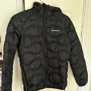 Svart dunjacka Peak Performance - Svart dunjacka från Peak Performance med vågformat quiltat mönster. Jackan har huva, hel dragkedja framtill och två sidofickor med dragkedja. Perfekt för kalla dagar och riktigt snygg till streetwear-stilen.