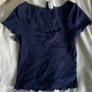 Mörkblå ribbad topp från H&M Divided - Mörkblå ribbad topp från H&M Divided med korta ärmar och söt spetskant vid urringningen. Toppen har tre knappar framtill och vågiga kanter vid ärmslut och nederkant. Perfekt för dig som gillar en enkel men ändå detaljrik stil.