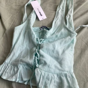 Turkos  linnetopp med snörning Gina Tricot - Supersöt turkos linnetopp från Gina Tricot i storlek XS. Toppen har snygg snörning framtill, volangkant nertill och är ärmlös. Perfekt för varma dagar och ger en fräsch vibe till din outfit. Kostar 300kr ny
