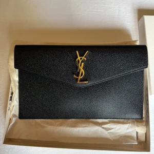 CLUTCH / kuvertväska från Saint Laurent - UPTOWN POUCH IN GRAIN DE POUDRE. I jättefint skick,  endast använt den 1 gång. Dustbag, certifikat, box osv.  27 X 16 X 2 CM. GOLD-TONED METAL HARDWARE MAGNETIC CLOSURE INTERIOR: ONE MAIN COMPARTMENT  100% CALFSKIN LEATHER.     Jag vill ej byta, bara sälja!😊