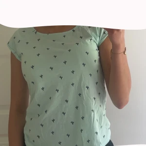 Mintgrön t-shirt med kaktusmönster Sinsay - Fräsch mintgrön t-shirt från Sinsay med små svarta kaktusmönster över hela. T-shirten har korta ärmar och är gjord i mjuk bomull, perfekt för en chill och avslappnad stil.