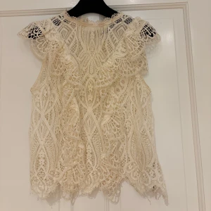 Vit spetsblus från Zara XS - Superfin vit blus från Zara i storlek XS, helt täckt av detaljerad spets och med volangdetaljer vid axlarna. Ärmlös modell med hög hals och vackert mönstrad spets som ger en romantisk vibe. Perfekt för dig som gillar unika toppar med mycket detaljer.