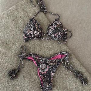 Svart och rosa bikini med paisleymönster - Snygg bikini med svart bas och paisleyinspirerat mönster i rosa, vitt och grönt. Triangeltopp med knyt i nacken och ryggen samt matchande trosa med knyt i sidorna och rosa foder. Perfekt för stranden eller poolen! HELT OANVÄND 🌟