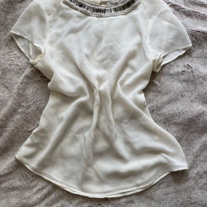 Vit blus med pärldetaljer från Zara - Supersnygg vit blus från Zara Trafaluc med korta ärmar och rund halsringning dekorerad med pärlor och silvriga detaljer. Tunn och flowy i ett skirt material, perfekt för att lyfta din look. Knäppning i nacken för extra detalj. Detaljerna på halsen är skadade och använda väll!