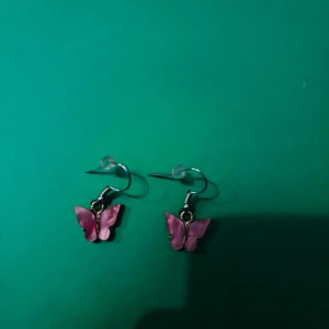 Rosa fjärilsörhängen i metall - Snygga örhängen med hängande fjärilar i rosa nyans. Fjärilarna har en glansig finish och är fästa på krokar i silverfärgad metall. Perfekta för att ge din look en söt och lekfull touch.
