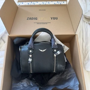 Zadig & Voltaire väska - Snygg svart handväska från Zadig & Voltaire i skinn med silverfärgade detaljer och dragkedjor framtill. Väskan har två handtag och ikonisk logga i silver på framsidan. Perfekt storlek och får plats med allt nödvändigt i. Lägg prisförslag vid intresse, tryck inte på köp nu!! Sällan använd och köpt på zadig och är i nyskick, nypris 5000 men säljer för 3500