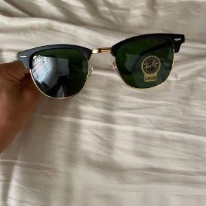 Ray-Ban Clubmaster svarta solglasögon - Snygga Ray-Ban Clubmaster solglasögon med klassisk svart ovandel och guldfärgade detaljer. Glasen är mörkgröna och modellen har en retroinspirerad halvbåge. Perfekta för dig som vill ha en tidlös och cool look.