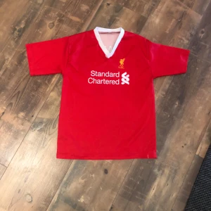 Liverpool Salah 11 röd fotbollströja - Röd Liverpool fotbollströja med vitt V-ringad krage, tryckt med Standard Chartered-logga på bröstet och M. Salah 11 på ryggen. Klassisk design med klubbmärke i gult på bröstet. Perfekt för fans av Mohamed Salah och Liverpool.