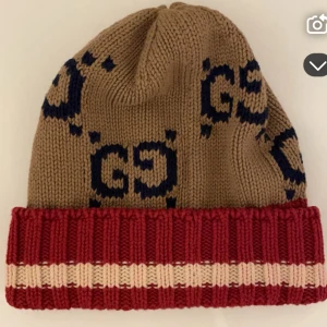 Gucci stickad mössa med logga - Snygg stickad mössa från Gucci i beige med mörkblå GG-loggor. Mössan har en bred röd uppvikt kant med en beige rand. Perfekt accessoar för att lyfta din vinterstil och hålla dig varm.