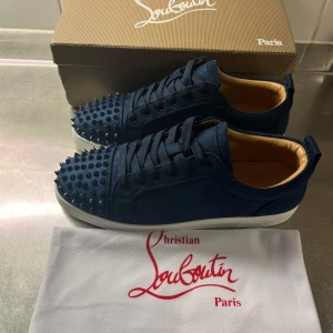 Mörkblå sneakers  - Snygga mörkblå sneakers  i mocka med coola nitar på tån och klassisk röd sula. Skorna har snörning och vit platt sula, samt lyxig innersida i skinn. Perfekta för dig som vill sticka ut med exklusiv streetstyle.