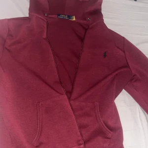 Vinröd hoodie från Polo Ralph Lauren - Säljer en vinröd hoodie från Polo Ralph Lauren med dragkedja och huva. Tröjan har klassisk Polo-logga broderad på bröstet, känguruficka framtill och långa ärmar. Perfekt för dig som gillar stilrena och bekväma plagg.