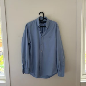 Blå skjorta från Ralph Lauren - Klassisk ljusblå skjorta från Ralph Lauren med knappar framtill och broderad logga på bröstet. Skjortan har lång ärm och normal passform, perfekt till jeans eller chinos. Tillverkad i mjuk bomull för en skön känsla hela dagen.Ny skick! Nypris runt 1200, pris kan diskuteras