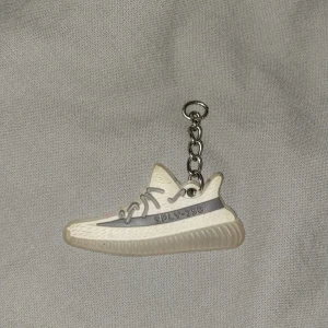 Adidas Yeezy Boost 350 nyckelring - Nyckelring i form av en Adidas Yeezy Boost 350 sneaker. Designen är detaljerad med grå snörning, grå rand med SPLY-350-tryck och en beige/vit ovandel. Perfekt accessoar för sneakerheads som vill visa sin stil även på nyckelknippan.