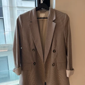Rutig kavaj från H&M i beige och svart - Snygg rutig kavaj från H&M i beige och svart med dubbelknäppning och klassisk krage. Kavajen har långa ärmar och två fickor framtill. Perfekt för dig som vill ha en stilren och trendig look.