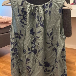 Grön blommig ärmlös linnetopp - Snygg ljusgrön ärmlös linnetopp med mörkblått blommönster. Toppen har en avslappnad passform och lätt rynk vid halsringningen för en luftig känsla. Perfekt för varma dagar och enkel att styla med jeans eller kjol.