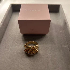 Guldig statementring från Edblad - Cool och chunky ring från Edblad i guldtonad metall. Ringen har flera rader med små konformade nitar som ger en edgy och unik look. Perfekt för dig som vill sticka ut med accessoarerna och gillar smycken som syns.       Ringen är i storlek 18.50