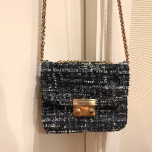 Svart och blå axelväska med kedja - Snygg axelväska i svart och blått med guldfärgad kedja. Väskan har ett  lås i guldmetall och ett innerfack med dragkedja. Det går att justera kedjan så man kan ha den både lång och kort. Låset har lite slitage men inget man tänker på. Säljer på grund av att den inte kommer till användning 💘