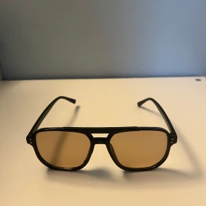 Svarta glasögon med bruna glas - Snygga solglasögon med svart pilotinspirerad båge och bruna tonade glas. Ramen är i plast och har en klassisk dubbla nitar vid sidorna. Perfekt accessoar för att lyfta din stil under soliga dagar.