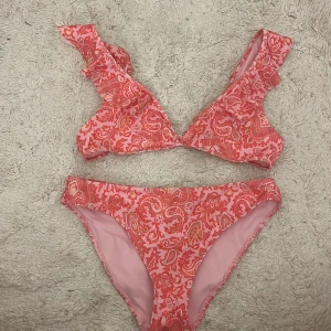 Rosa bikini med volang och paisleymönster - Superfin bikini i rosa med rött paisleymönster och volangdetaljer på axelbanden. Överdelen har breda axelband och knäppe bak, medan underdelen är klassisk med medelhög midja. Perfekt för stranden eller poolen!