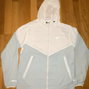 Nike vindjacka i vitt och ljusblått - Nike vindjacka med huva i vitt och ljusblått. Jackan har dragkedja framtill, två fickor med vita detaljer och reflekterande detaljer på ärmarna. Perfekt för blåsiga dagar och sportiga outfits. Klassisk Nike-logga på bröstet och justerbar huva.