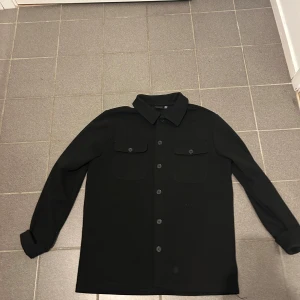 Svart overshirt med knappar - Stilren svart overshirt med klassisk krage och långa ärmar. Skjortan har två bröstfickor med lock och knappar framtill. Perfekt att bära öppen eller stängd för en clean look.