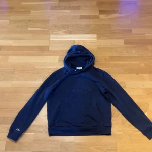 Marinblå hoodie från Lacoste - Snygg marinblå hoodie från Lacoste med broderad logga och krokodil på bröstet. Tröjan har huva med snörning, känguruficka och långa ärmar. Perfekt för dig som gillar stilren och sportig look.