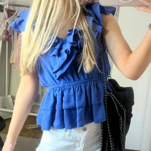 Blå volangblus med v-ringning - Supersöt blå blus med volanger längs v-ringningen och axlarna. Toppen är ärmlös och har resår i midjan för en snygg passform. Perfekt att styla med jeans eller kjol för en trendig look. Materialet känns lätt och luftigt.
