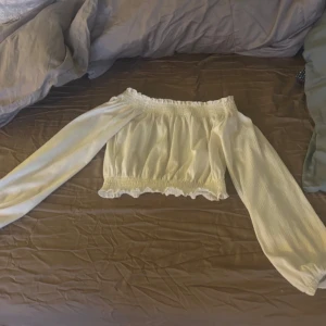 Offshoulder ljusgul blus med volang - Superfin offshoulder blus i vit färg med långa ärmar och smockad resår upptill och nertill. Volangdetaljer vid kanterna ger en söt och trendig look. Perfekt till jeans eller kjol för en fräsch stil. 