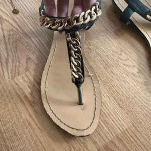 Svarta sandaler med guldkedja - Snygga svarta sandaler med bred guldfärgad kedjedetalj över foten och längs remmen. Platt sula och öppen tå, perfekt för varma dagar. Stilsäkra och lätta att matcha till sommarens outfits