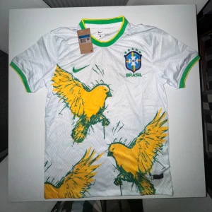 Nike x Brasilien fotbollströja  - Snygg vit Brasilien fotbollströja från Nike med gröna och gula detaljer på krage och ärmslut. Framsidan har stora gula fåglar i grafisk stil och Brasilien-emblem på bröstet. Tillverkad i Dri-FIT material som andas, perfekt för match eller träning.
