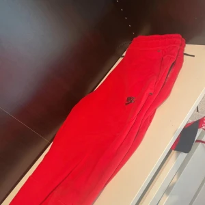 Röda mjukisbyxor från Nike - Snygga röda mjukisbyxor från Nike med svart logga på benet. Byxorna har elastisk midja med svart snörning och är tillverkade i mjukt bomullsmaterial. Perfekta för chill eller träning, med en loose fit och klassisk sportig look.