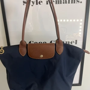 Marinblå Longchamp handväska - Snygg marinblå handväska från Longchamp med bruna skinnhandtag och lock. Väskan har guldiga detaljer, dragkedja upptill och ett rymligt huvudfack med innerficka. Perfekt storlek för att få plats med allt du behöver under dagen.