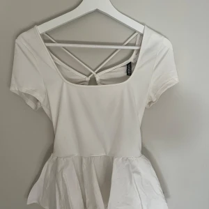 Vit peplumtop med snörning H&M Divided - Superfin vit peplumtop från H&M Divided med korta ärmar och snygg snörning i ryggen. Toppen har en öppen rygg med knyt och criss-cross-detaljer, samt en volangig nederdel. Perfekt för dig som gillar trendiga och feminina plagg.