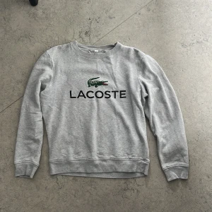Grå sweatshirt från Lacoste - Snygg grå sweatshirt från Lacoste med klassisk krokodil-logga och stor svart text på bröstet. Tröjan har rund hals, långa ärmar och ribbade muddar. Materialet är mjuk bomullsmix som känns skönt mot huden.
