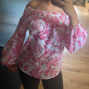 Offshoulder blommig blus i rosa och vitt - Superfin offshoulder blus med stora rosa blommor på vit botten. Vida ärmar med resår vid handlederna och smock upptill som håller blusen på plats. Luftigt material, perfekt för varma dagar. Snygg till både jeans och kjol!