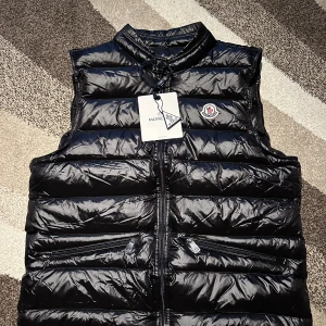 Moncler väst  - Säljer denna snygga och klassiska Moncler-väst i svart, storlek M. Västen är helt ny med tag kvar, aldrig använd. Fick en exakt likadan i present av en släkting, så denna behöver jag inte.  Modellen är tidlös, dunfylld och perfekt både som ytterplagg vid mildare väder eller under jackan för extra värme. Västen har två dragkedjefickor framtill och ikoniska Moncler-loggan på bröstet.