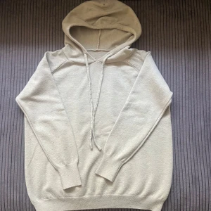 Beige hoodie med snörning i merinoull - En stilren beige hoodie med långa ärmar och huva. Tröjan har snörning framtill och är tillverkad i ett mjukt, stickat material. Perfekt för dig som gillar en enkel och clean look. Passformen är normal och muddar finns vid ärmslut och nederkant. Storlek S herr 