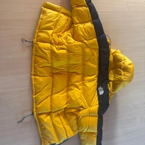 Gul och svart dunjacka The North Face - Säljer en ikonisk gul och svart dunjacka från The North Face i storlek M. Jackan har huva, stora quiltade paneler och dragsko i nederkant. Perfekt för kalla dagar och riktigt snygg streetstil. Klassisk logga broderad på bröstet. Används typ 4 ggr.