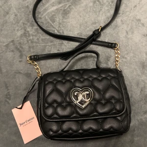Svart quiltad axelväska Juicy Couture - Supersöt svart axelväska från Juicy Couture med quiltat hjärtmönster och guldfärgade detaljer. Väskan har ett hjärtformat JC-spänne framtill, justerbar axelrem med kedja och tryckknapp. Insidan är fodrad med logotyp-tyg i svartvitt.