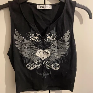 Svart croppad linnetopp FB Sister - Svart croppad linnetopp från FB Sister med ett stort tryck framtill av ett hjärta med vingar och silvriga detaljer. Toppen är ärmlös och har en liten v-ringning. Ribbad struktur i tyget ger en cool känsla.