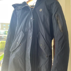 Parkas från 8848 Altitude med fuskpäls - Mörkblå parkas från 8848 Altitude med mysig fuskpäls på huvan och quiltat innerfoder. Jackan har stora fickor med knappar, dragkedja och knappstängning framtill samt en snygg detalj med logga på bröstet. Perfekt för kalla dagar när du vill hålla stilen.