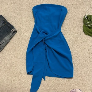 Blå axelbandslös kortklänning - Snygg blå axelbandslös kortklänning med knyt framtill som ger en cool twist. Klänningen har en figurnära passform och är tillverkad i ett mjukt tyg som känns skönt mot huden. Perfekt för dig som vill sticka ut med en färgstark look. Man knyter själv