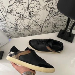 Burberry sneakers med rutigt mönster - Snygga svarta sneakers från Burberry med klassiskt rutigt mönster i beige, svart och brunt på sidorna och hälen. Skorna har snörning, platt sula i ljus färg och detaljer i skinn och mocka. Perfekta för dig som vill ha en stilren men ändå unik look. Lite smutsiga men går att rengöra och de är storlek 44 men sitter som 45