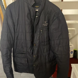 Svart quiltad jacka Aeronautica Militare - Snygg svart quiltad jacka från Aeronautica Militare med diskret logga på bröstet och dragkedjeficka. Jackan har knappar framtill och en stilren, rak passform. Perfekt för dig som gillar en clean och sportig look.